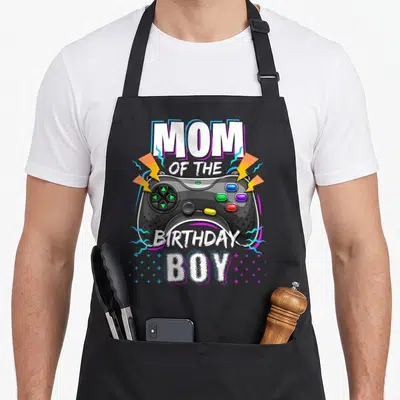 Mum birthday apron - Game controller graphic with MOM OF THE BIRTHDAY BOY text and lightning bolts — shown on a black cotton canvas apron against a white background