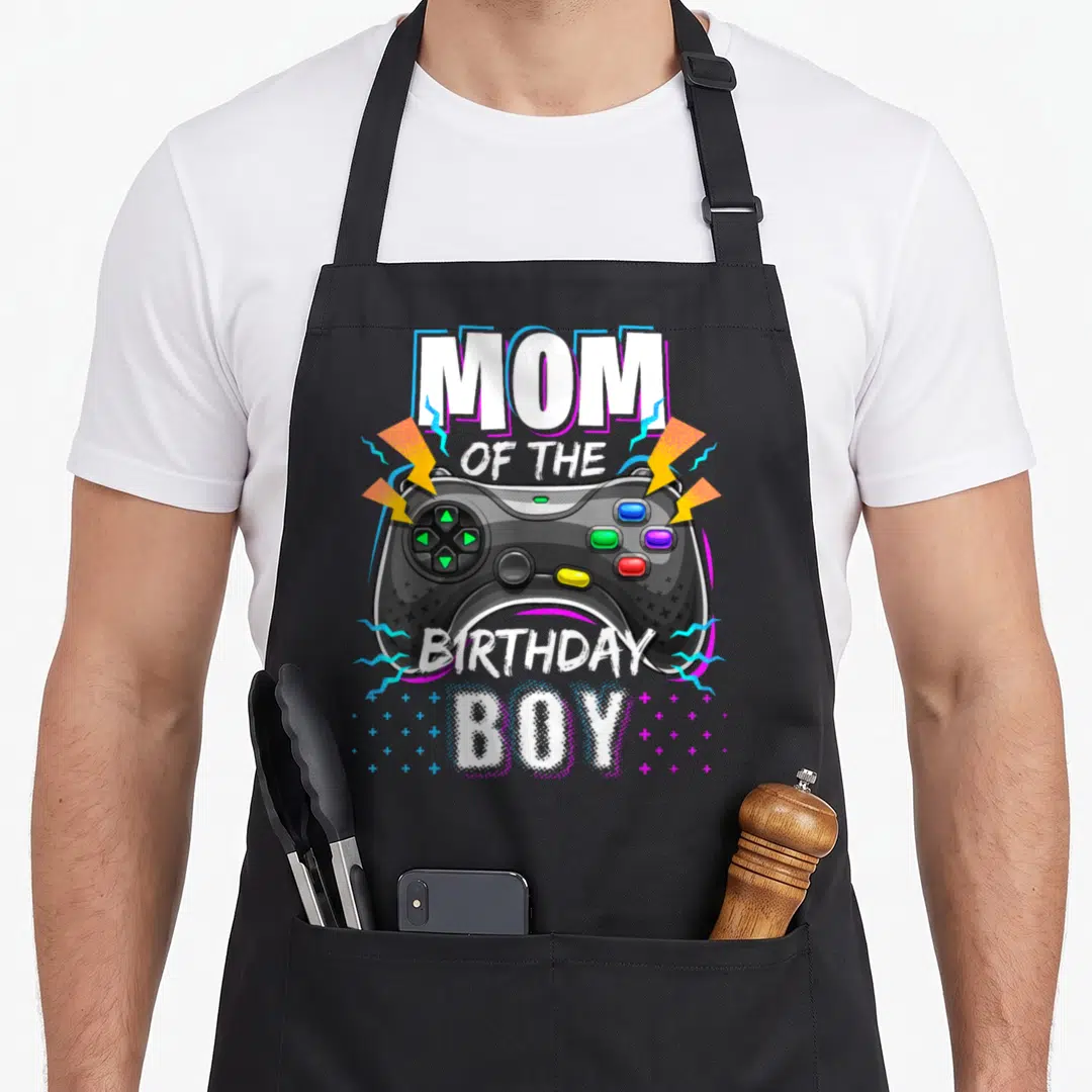 Mum birthday apron - Game controller graphic with MOM OF THE BIRTHDAY BOY text and lightning bolts — shown on a black cotton canvas apron against a white background
