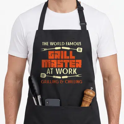 Chilling grill master apron - Orange and beige distressed text reading THE WORLD FAMOUS GRILL MASTER AT WORK GRILLING & — shown on a black cotton canvas apron against a white background