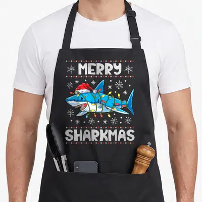Sharkmas xmas apron - Cartoon blue shark in red Santa hat wrapped in lights with MERRY SHARKMAS text and snowflakes — shown on a black cotton canvas apron against a white background