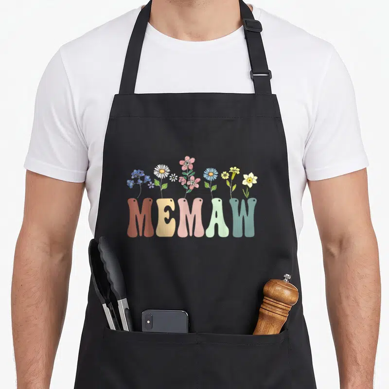 Memaw floral apron - Cartoon wildflowers in pink blue yellow green and orange growing from bold MEMAW letters — shown on a black cotton canvas apron against a white background