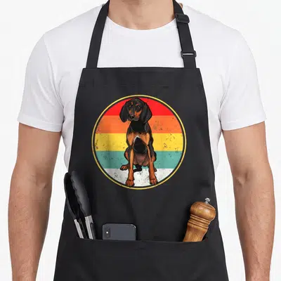 Sunset coonhound dog apron - Seated black and tan hound in front of circular retro sunset stripes — shown on a black cotton canvas apron against a white background