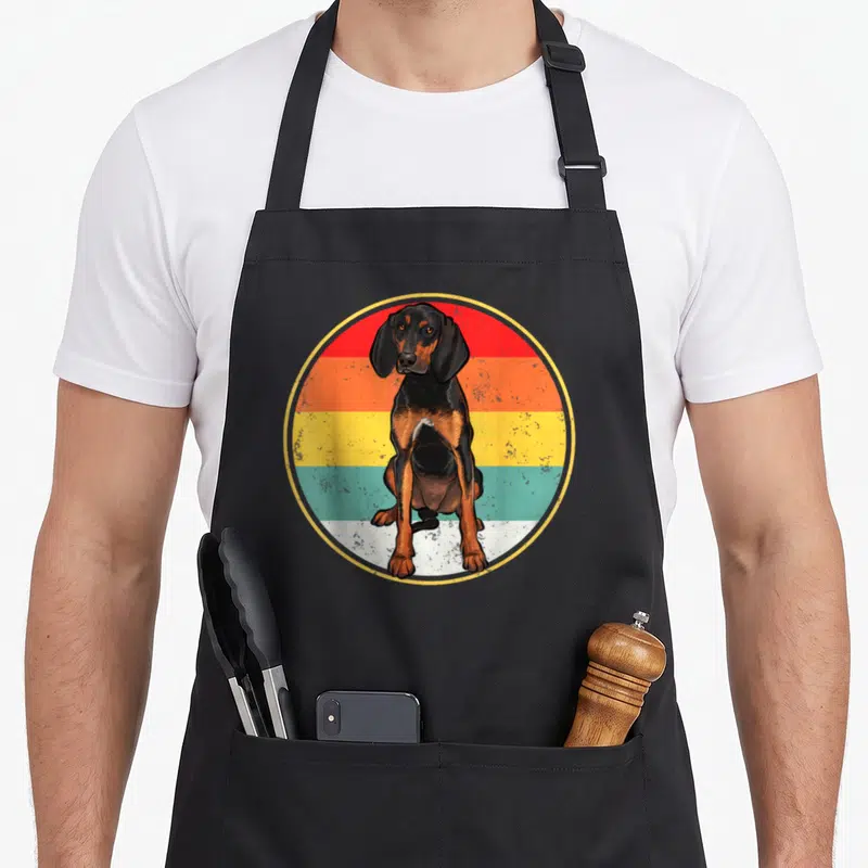 Sunset coonhound dog apron - Seated black and tan hound in front of circular retro sunset stripes — shown on a black cotton canvas apron against a white background