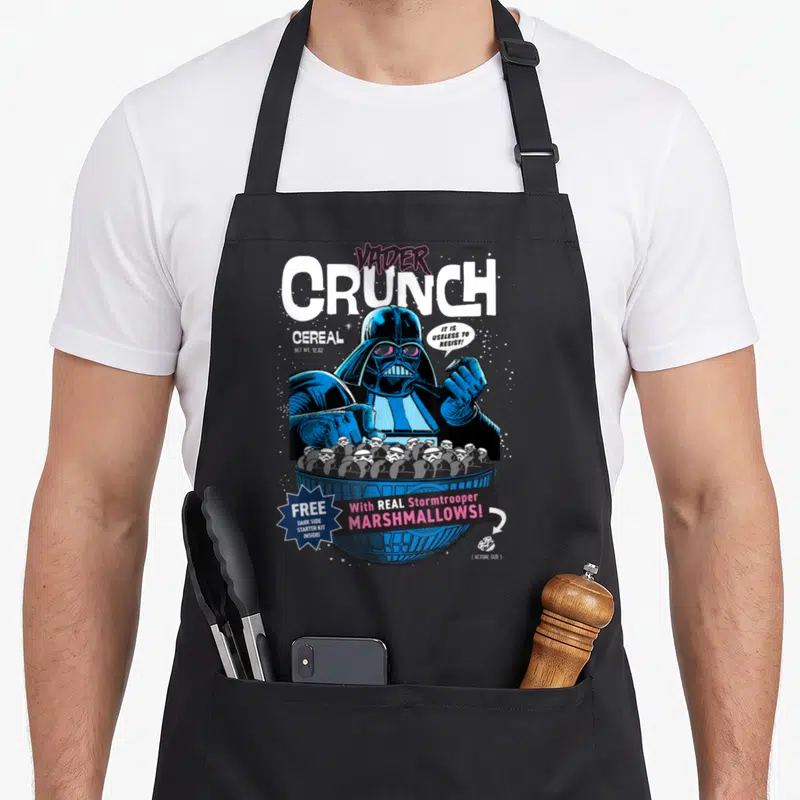 Vader cereal apron - Darth Vader cartoon holding bowl of stormtrooper marshmallows with retro cereal box text — shown on a black cotton canvas apron against a white background