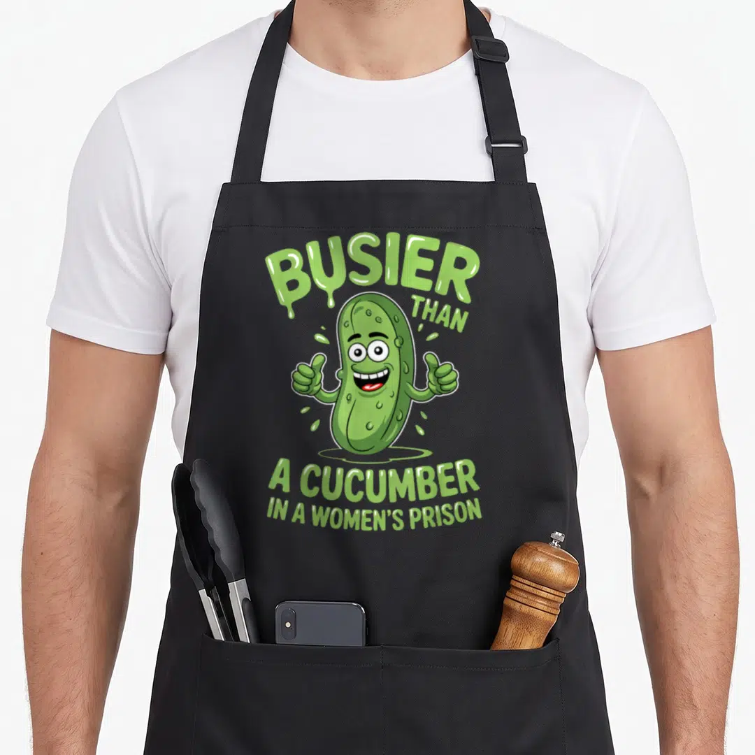 Busier cucumber apron - Green cartoon cucumber character giving two thumbs up with arched and stacked green text — shown on a black cotton canvas apron against a white background