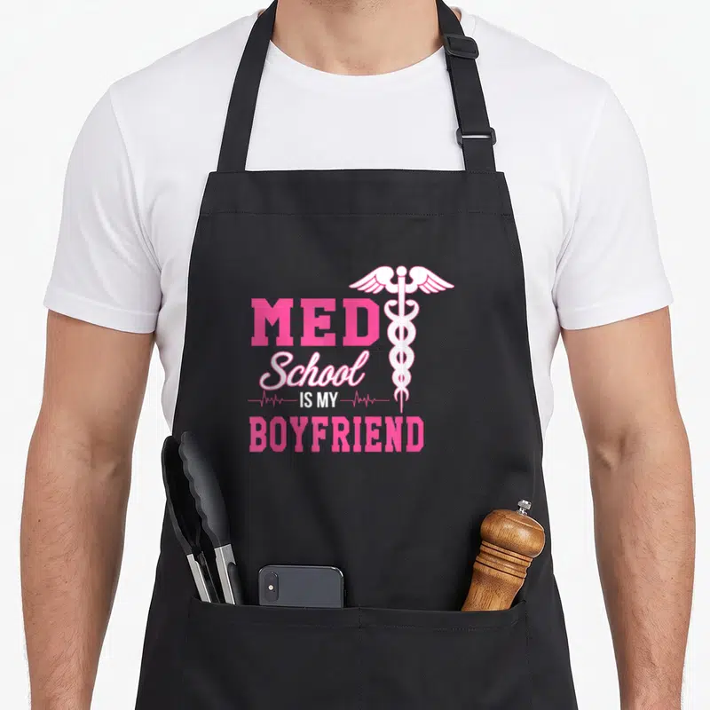Boyfriend doctor apron - Pink and white Med School Is My Boyfriend text with caduceus symbol — shown on a black cotton canvas apron against a white background