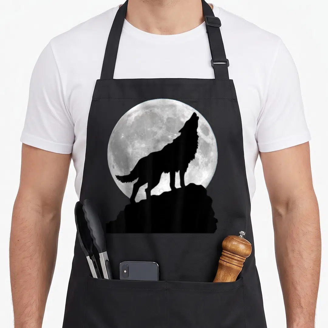 Wolf dog apron - Black wolf silhouette howling on a rock in front of a large grey and white full moon — shown on a black cotton canvas apron against a white background