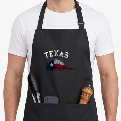 Funny armadillo texas flag apron - Cartoon armadillo with blue white and red flag coloring under arched white TEXAS text — shown on a black cotton canvas apron against a white background