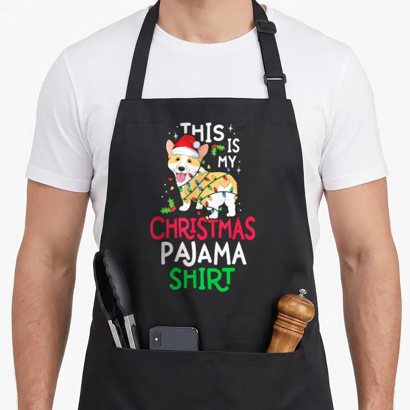 Pyjama christmas tree apron - Corgi in Santa hat wrapped in colored lights with text THIS IS MY CHRISTMAS PAJAMA SHIRT — shown on a black cotton canvas apron against a white background