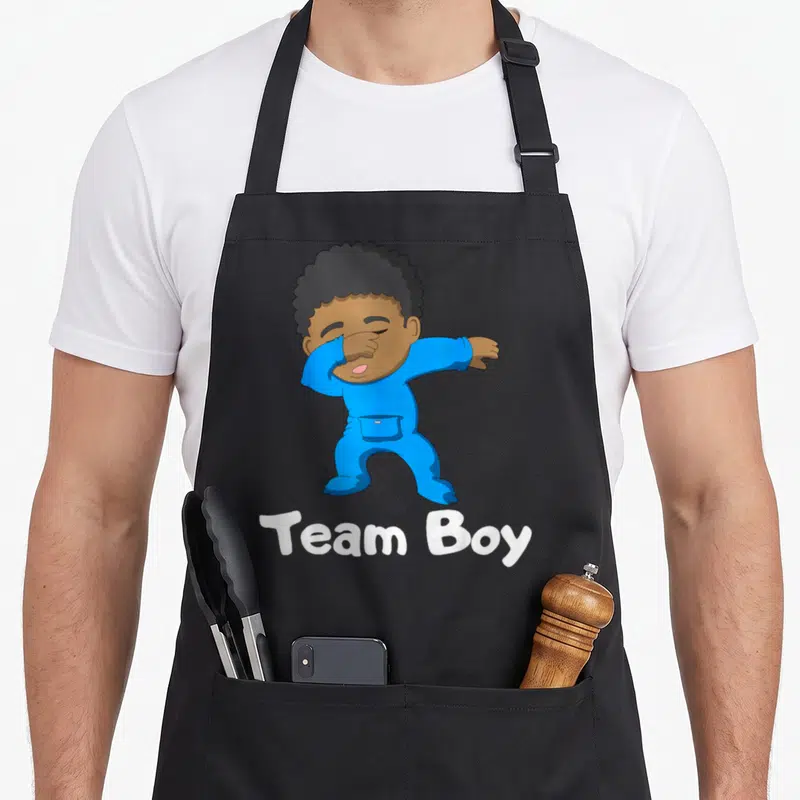 Gender dabbing boys apron - Cartoon baby in blue onesie dabbing with white Team Boy text underneath — shown on a black cotton canvas apron against a white background