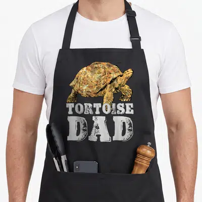 Tortoise african apron - Distressed tortoise illustration above white stacked text reading TORTOISE DAD — shown on a black cotton canvas apron against a white background
