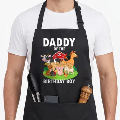 Daddy farm aprons - Daddy of the Birthday Boy text over cartoon farm animals, barn, and fence in white red green — shown on a black cotton canvas apron against a white background