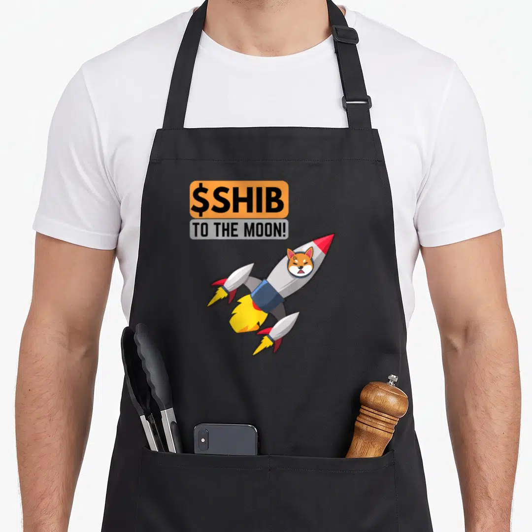 Shib shiba inu apron - $SHIB TO THE MOON! text above cartoon rocket with dog head in orange and grey — shown on a black cotton canvas apron against a white background