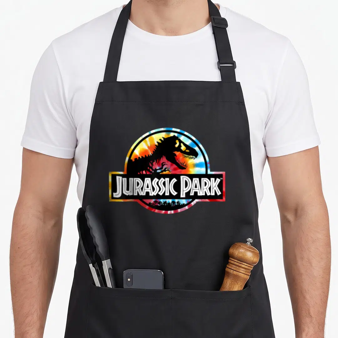 Circle tie dye apron - T-Rex skeleton in tie-dye circle with Jurassic Park text banner — shown on a black cotton canvas apron against a white background