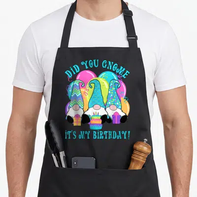 Gnome balloon apron - Three gnomes with patterned hats under DID YOU GNOME text with cake and presents — shown on a black cotton canvas apron against a white background