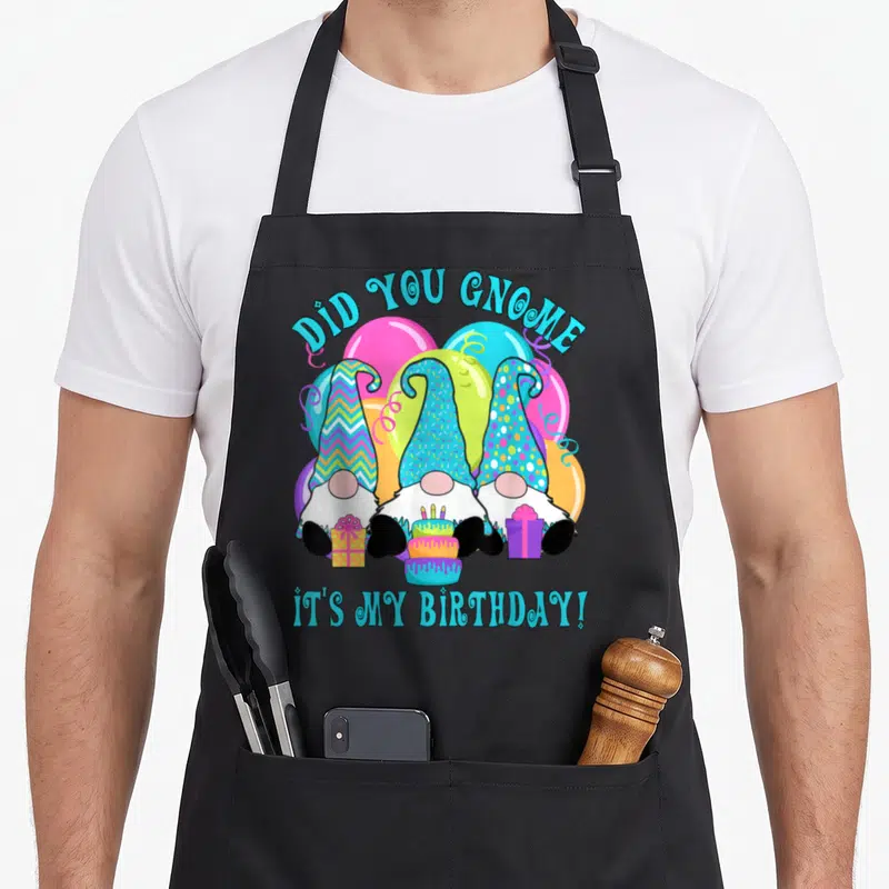 Gnome balloon apron - Three gnomes with patterned hats under DID YOU GNOME text with cake and presents — shown on a black cotton canvas apron against a white background