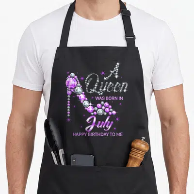 Funny july baking queen apron - Purple diamond high heel shoe next to A Queen Was Born In July Happy Birthday To Me text — shown on a black cotton canvas apron against a white background