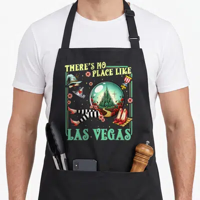 Brick road apron - There's No Place Like Las Vegas text surrounding a snow globe with castle, witch hat, and striped — shown on a black cotton canvas apron against a white background