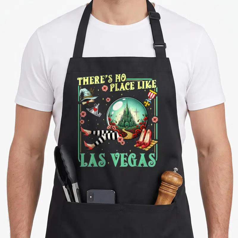 Brick road apron - There's No Place Like Las Vegas text surrounding a snow globe with castle, witch hat, and striped — shown on a black cotton canvas apron against a white background