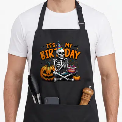 Skeleton funny halloween apron - Cartoon skeleton holding birthday cake between orange pumpkins with arched text — shown on a black cotton canvas apron against a white background