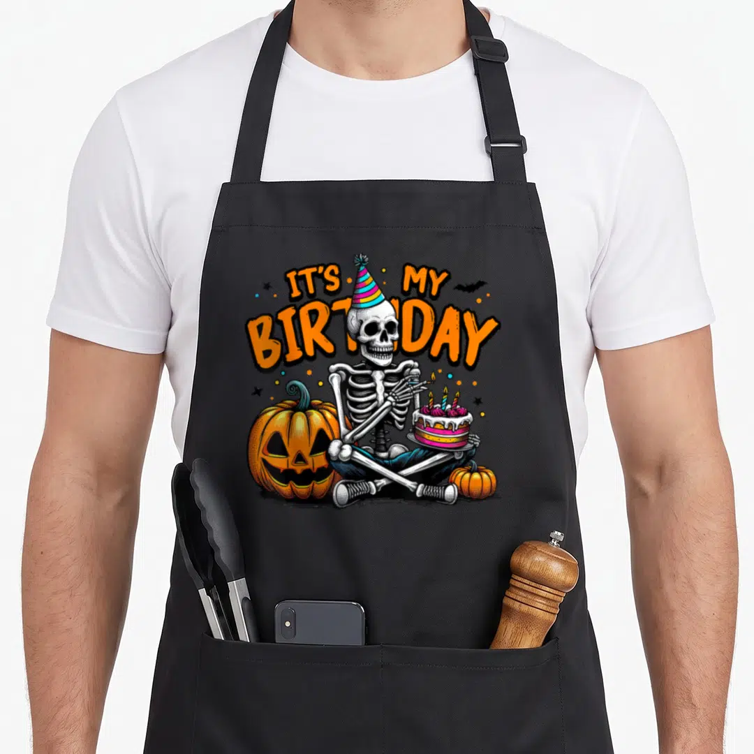 Skeleton funny halloween apron - Cartoon skeleton holding birthday cake between orange pumpkins with arched text — shown on a black cotton canvas apron against a white background