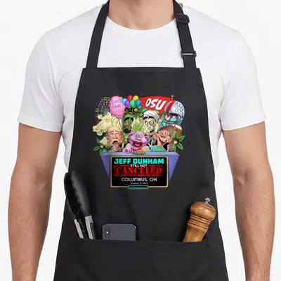 Jeff dunham hollywood apron - Puppets clustered behind blue stage with OSU banner and gold plaque reading still not — shown on a black cotton canvas apron against a white background