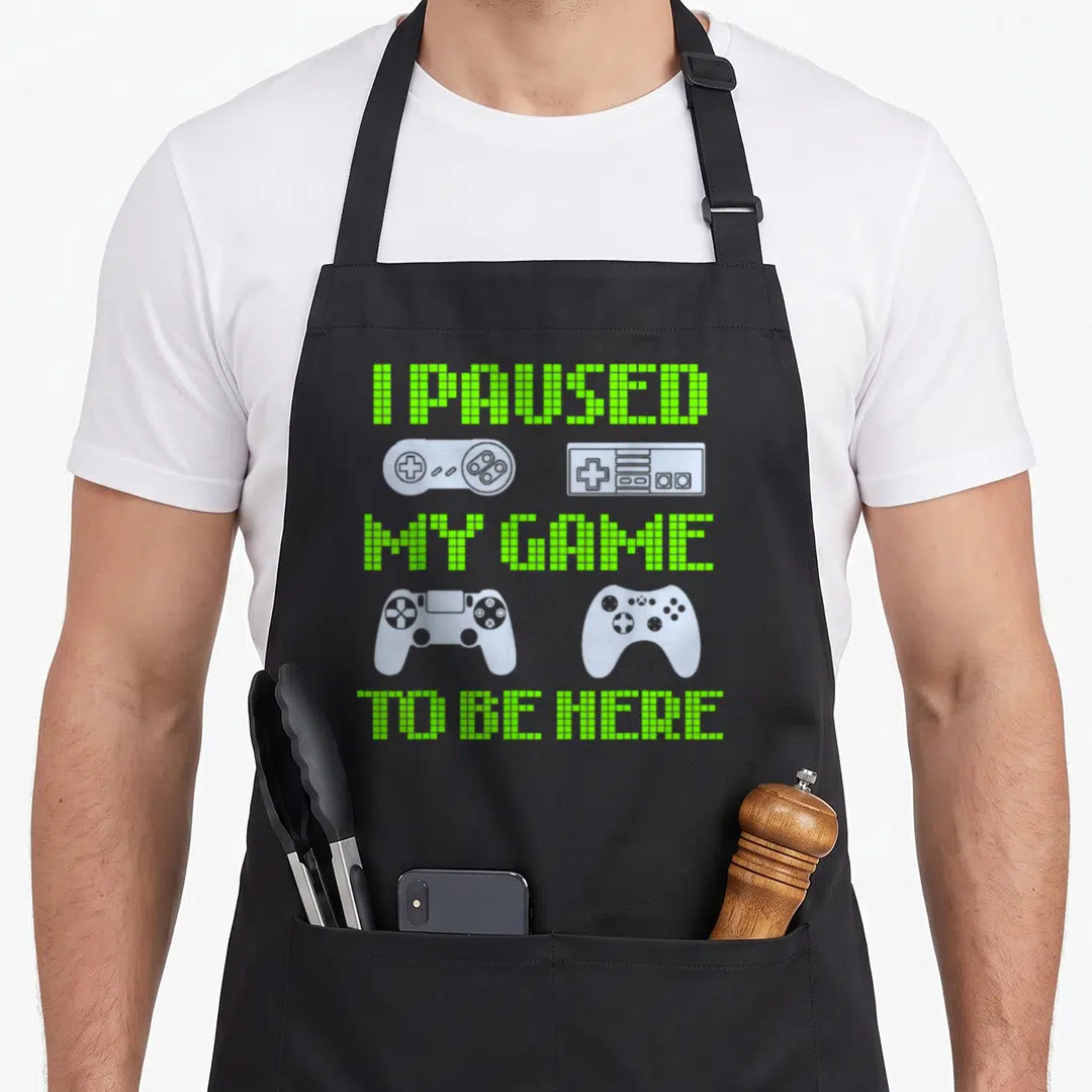 Vintage paused video game apron - Lime green pixel text reading I PAUSED MY GAME TO BE HERE flanked by grey retro — shown on a black cotton canvas apron against a white background
