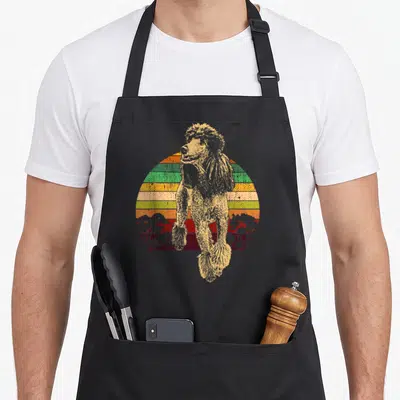 Miniature standard size of apron - Beige poodle standing in front of a striped semi-circle sun with red tree — shown on a black cotton canvas apron against a white background