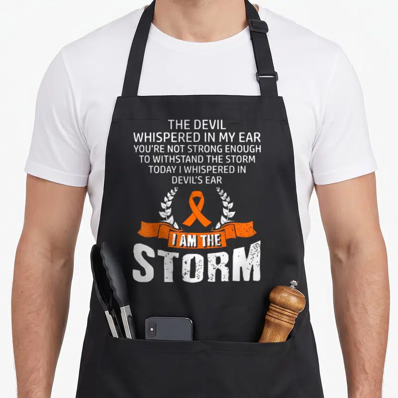 Storm leukemia awareness apron - White stacked text and orange ribbon inside white laurel wreath on black background — shown on a black cotton canvas apron against a white background