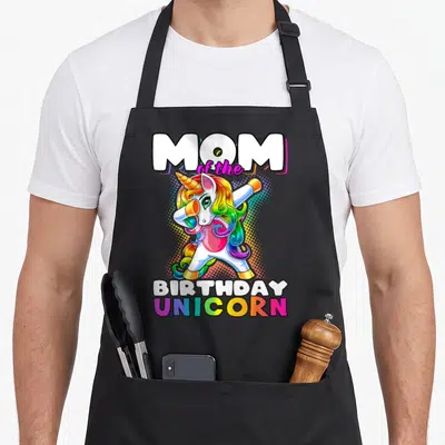 Birthday unicorn family aprons - Dabbing unicorn with rainbow mane and MOM of the BIRTHDAY UNICORN text — shown on a black cotton canvas apron against a white background