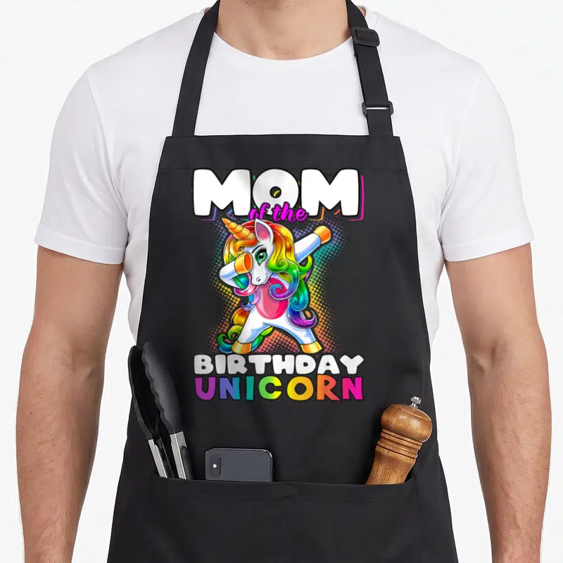 Birthday unicorn family aprons - Dabbing unicorn with rainbow mane and MOM of the BIRTHDAY UNICORN text — shown on a black cotton canvas apron against a white background