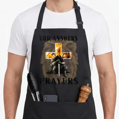 God prayer apron - Kneeling knight, orange cross, lion face, black text GOD ANSWERS PRAYERS — shown on a black cotton canvas apron against a white background