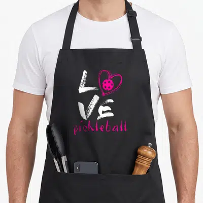 Cute pickleball apron - Vertical distressed white LOVE text with pink heart containing ball and pink script pickleball — shown on a black cotton canvas apron against a white background