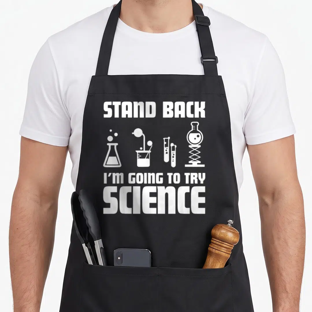 Stand chemistry apron - White text STAND BACK I'M GOING TO TRY SCIENCE above row of flasks and test tubes — shown on a black cotton canvas apron against a white background