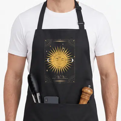 Dark moon gothic apron - Golden sun with face flanked by two crescent moons inside a starry rectangular border — shown on a black cotton canvas apron against a white background