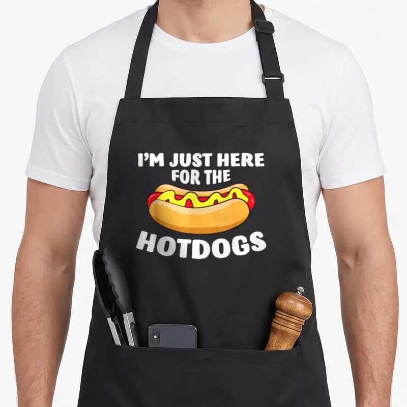 Funny hot dog apron - Cartoon hot dog with mustard and ketchup surrounded by white text I'm just here for the hotdogs — shown on a black cotton canvas apron against a white background