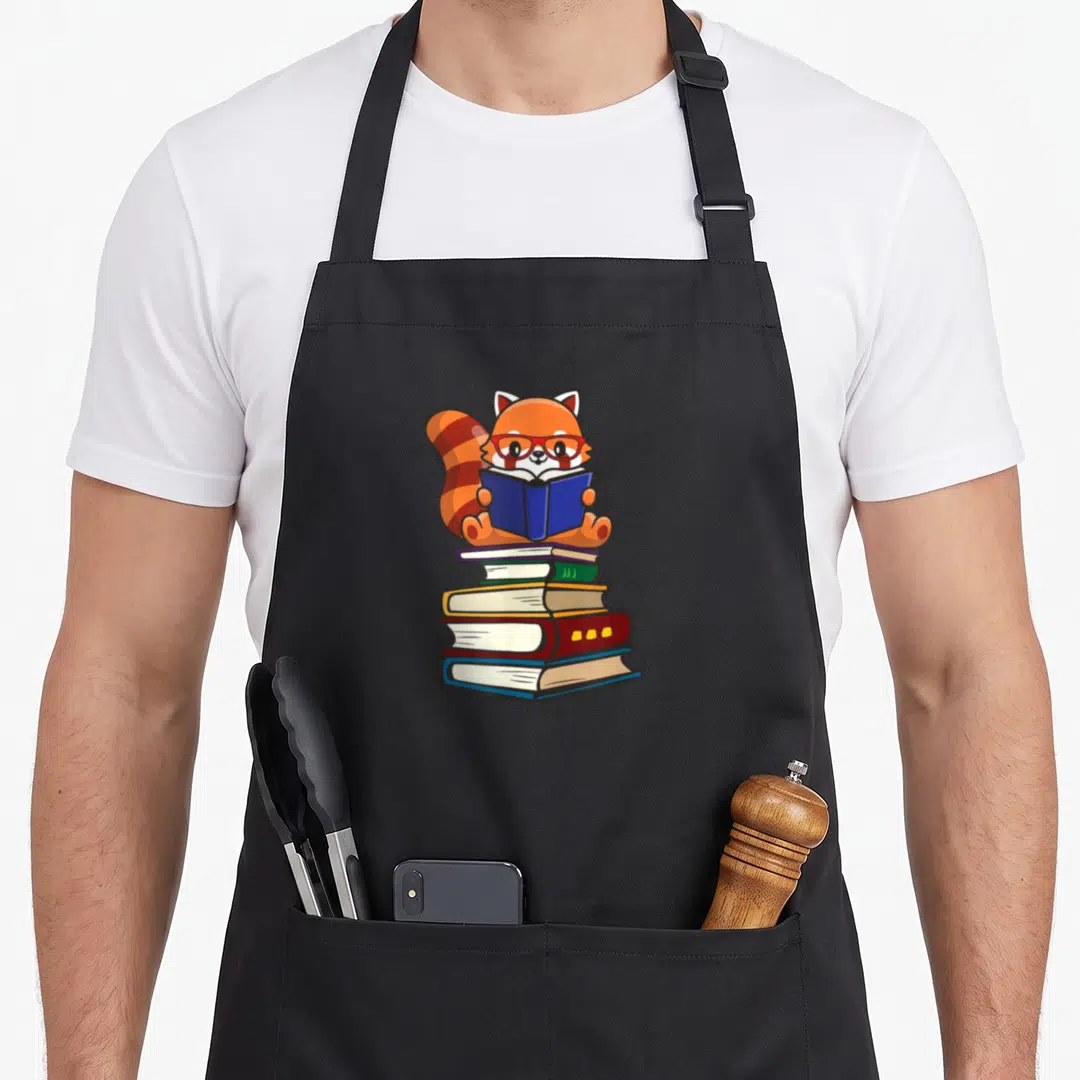 Reading kawaii apron - Orange red panda with red glasses holding blue book on stack of five colorful volumes — shown on a black cotton canvas apron against a white background