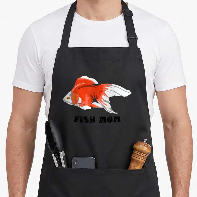 Fish silly apron - Orange and white cartoon goldfish above black FISH MOM text — shown on a black cotton canvas apron against a white background