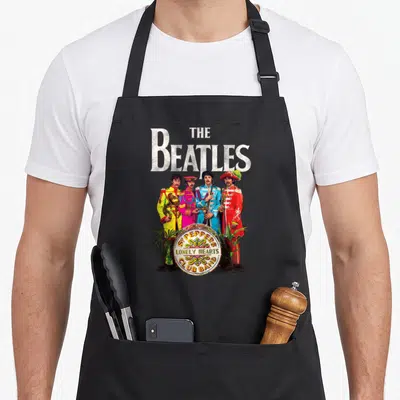Lonely sergeant apron - Four Beatles in colorful uniforms standing behind a drum with plants and album title text — shown on a black cotton canvas apron against a white background