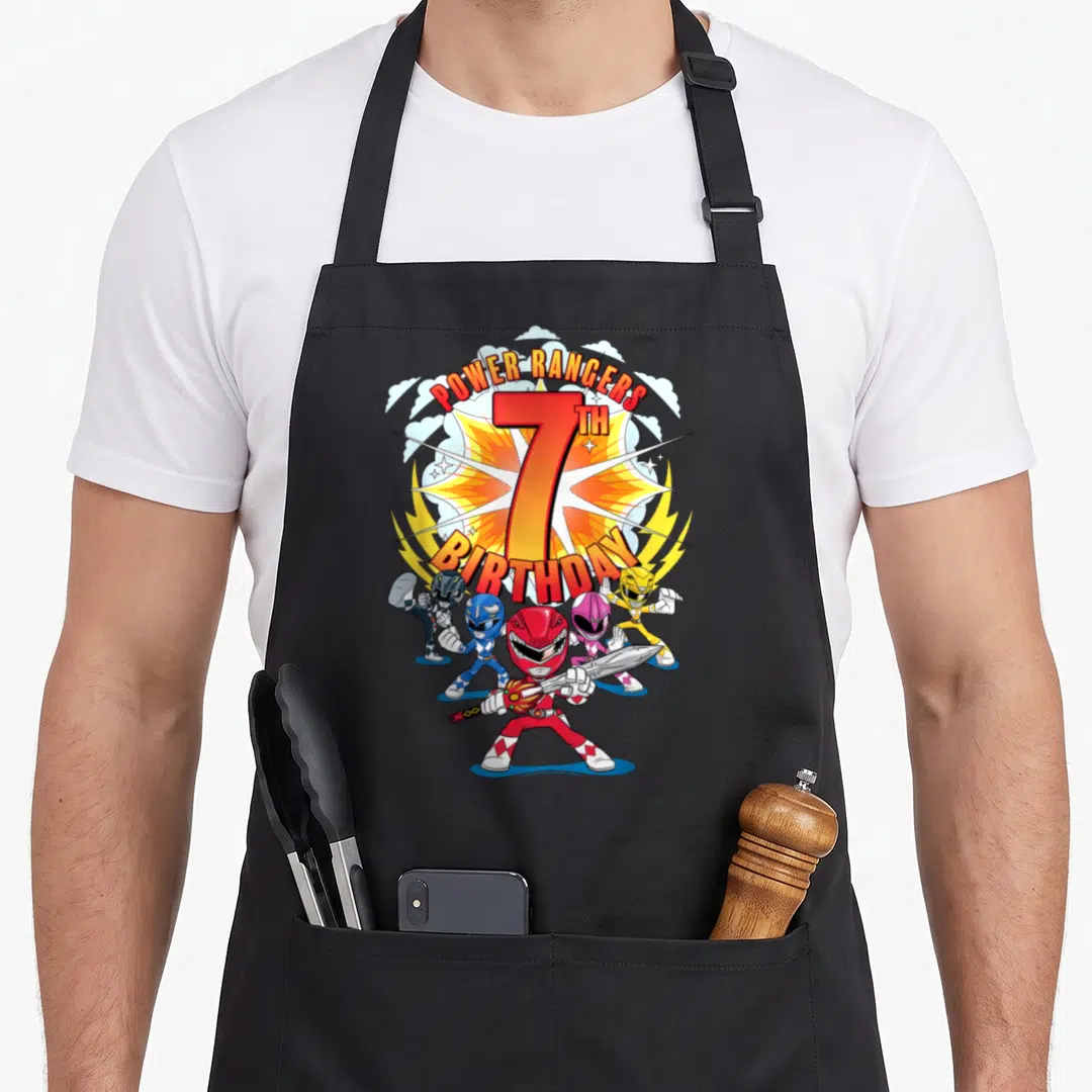 7th action shot birthday apron - Five Power Rangers in red yellow blue pink with arched text and large 7TH BIRTHDAY — shown on a black cotton canvas apron against a white background