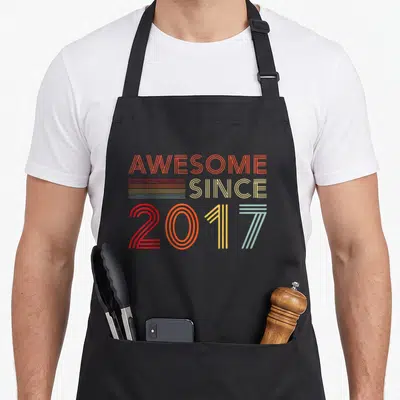 Decoration dad and son aprons - Stacked retro typography reading AWESOME SINCE 2017 in orange yellow red and blue-grey — shown on a black cotton canvas apron against a white background