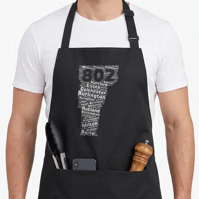 Authentic vermont apron - White text map of Vermont towns forming state shape on black apron — shown on a black cotton canvas apron against a white background