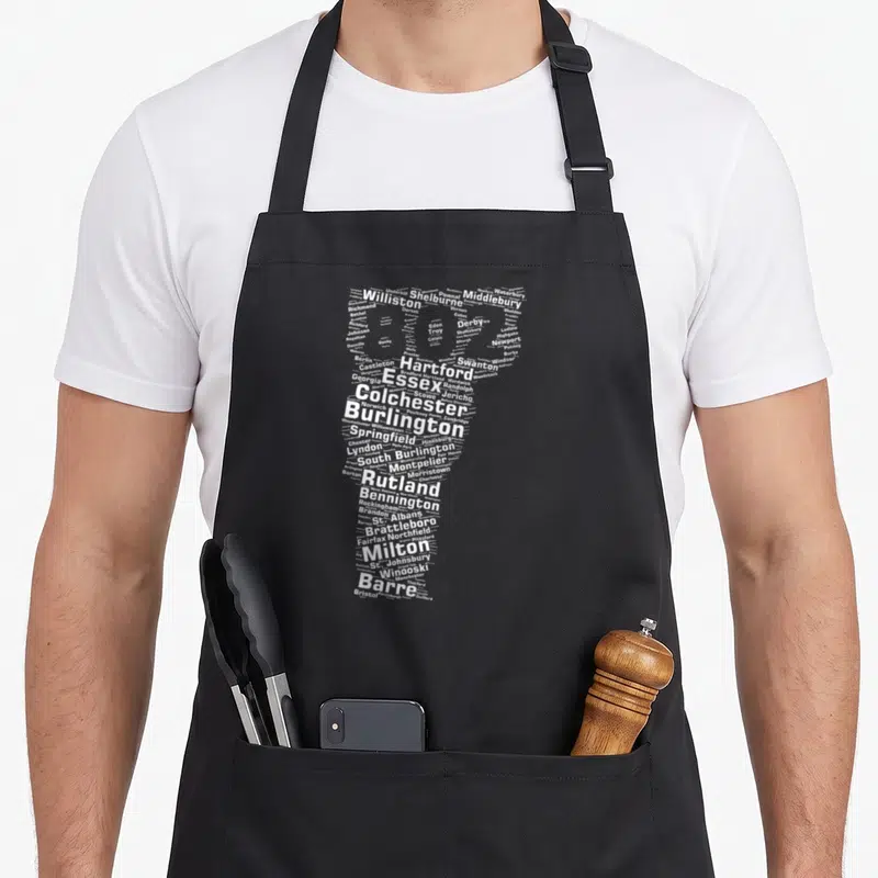 Authentic vermont apron - White text map of Vermont towns forming state shape on black apron — shown on a black cotton canvas apron against a white background