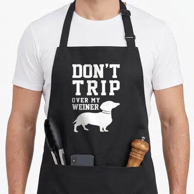 Trip funny dog apron - White stacked text reading Don't Trip Over My Weiner above a dachshund silhouette — shown on a black cotton canvas apron against a white background