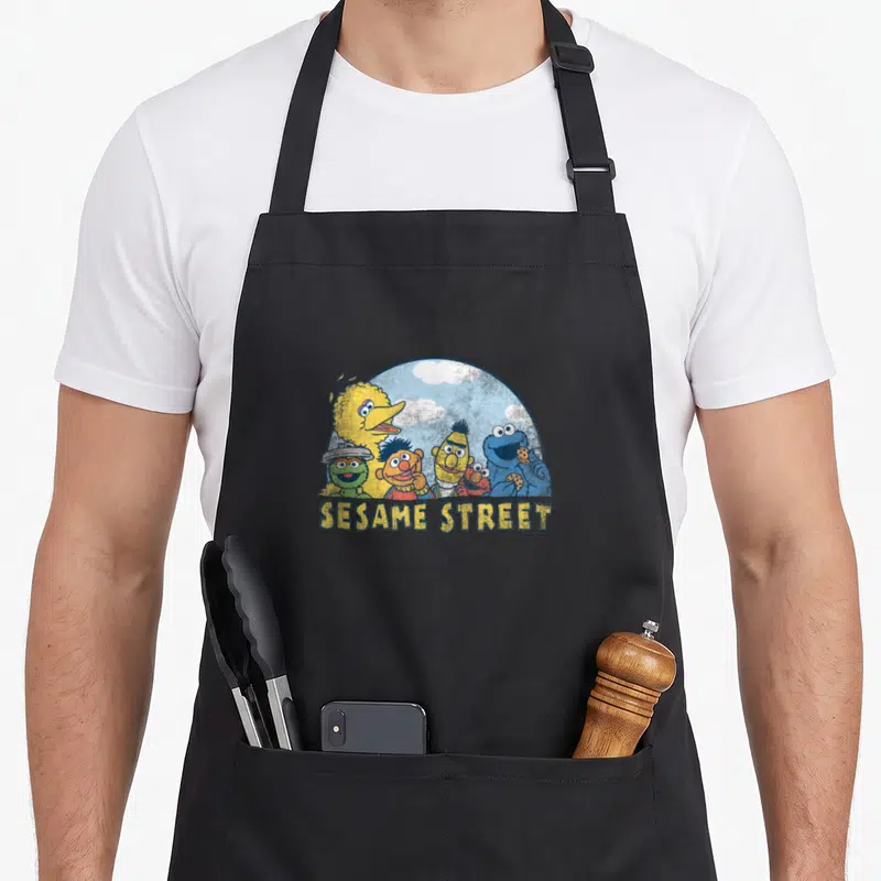 Vintage street apron - Sesame Street characters clustered in a blue semi-circle with clouds and yellow text below — shown on a black cotton canvas apron against a white background