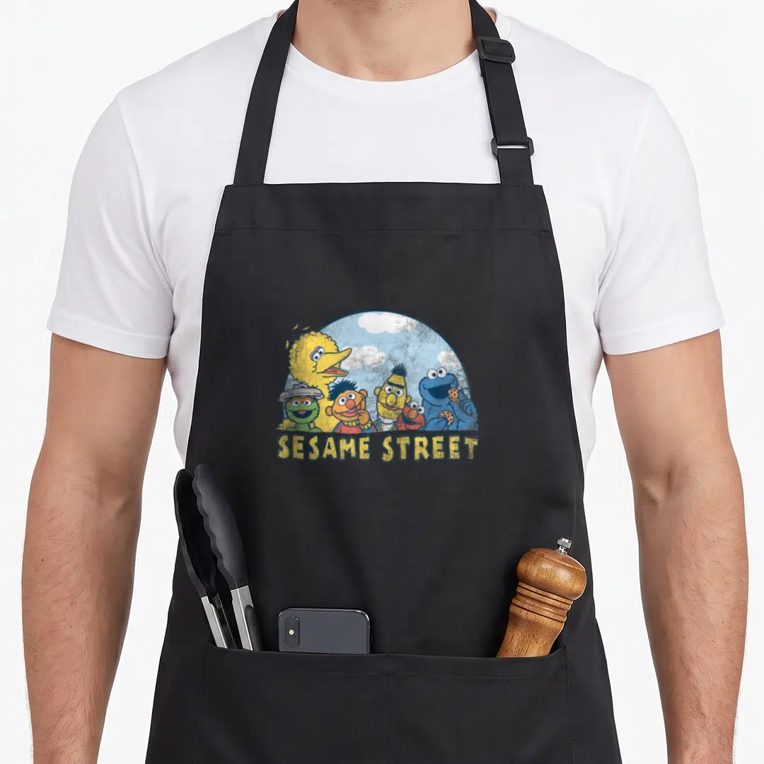 Vintage street apron - Sesame Street characters clustered in a blue semi-circle with clouds and yellow text below — shown on a black cotton canvas apron against a white background