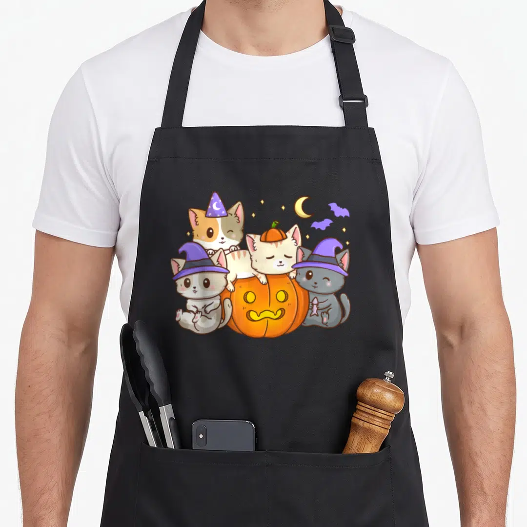 Halloween kawaii cooking apron - Three cats in purple witch hats clustering around an orange jack-o-lantern pumpkin — shown on a black cotton canvas apron against a white background