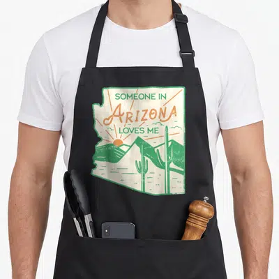 Someone arizona apron - Retro Arizona state outline with cacti, mountains, and rising sun in green and orange — shown on a black cotton canvas apron against a white background