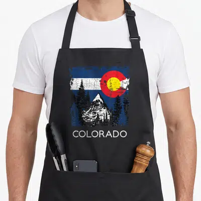 Flag mountain colorado apron - Distressed Colorado flag background with mountain peak, pine trees, and white text — shown on a black cotton canvas apron against a white background