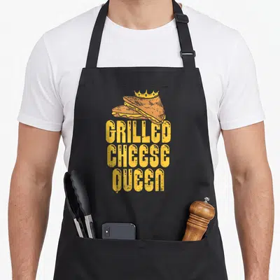 Queen maker apron - Crowned grilled cheese sandwich above distressed yellow stacked text reading GRILLED CHEESE QUEEN — shown on a black cotton canvas apron against a white background